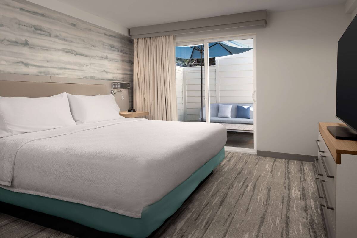 A hotel room with a large bed, a TV, a nightstand, and a view of a balcony with a blue umbrella.