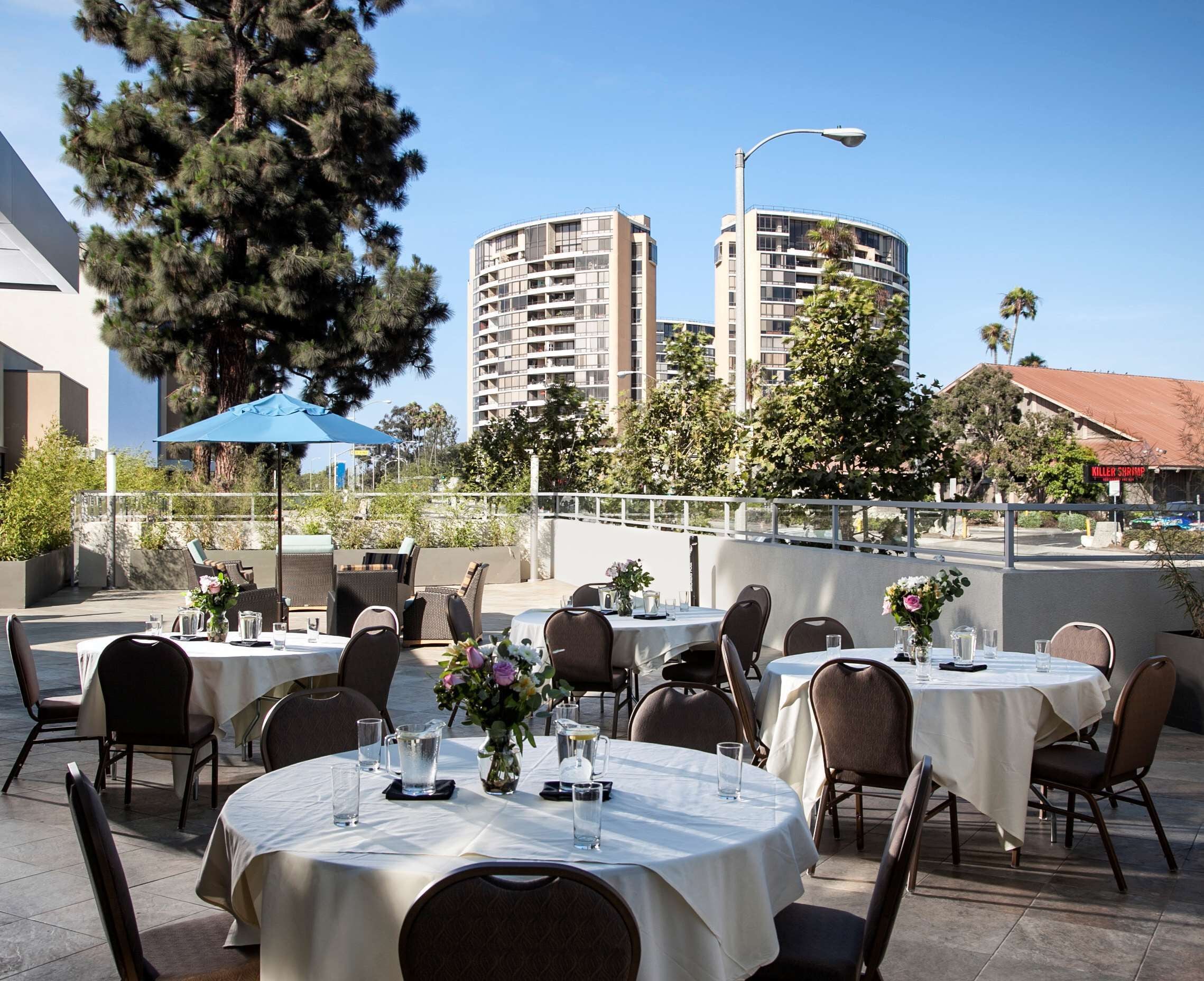 Un área de comedor al aire libre con mesas y sillas redondas, una sombrilla azul y vistas de edificios altos al fondo.