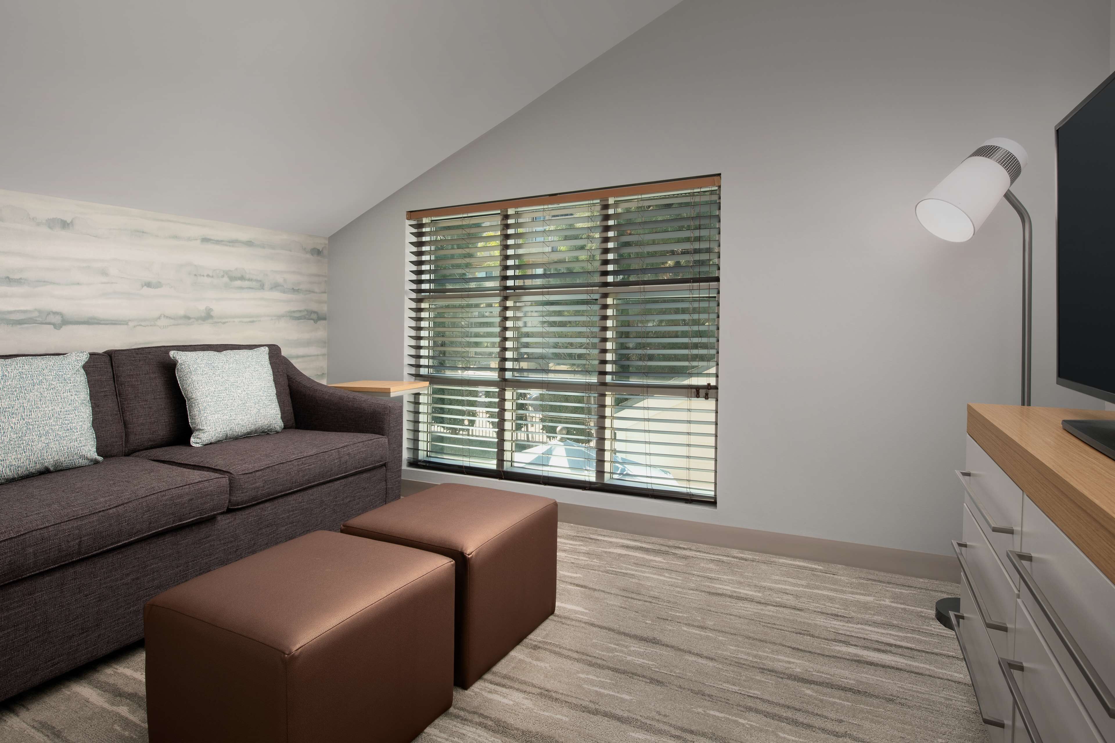 A modern living room with a sofa, ottoman, and a large window with blinds.