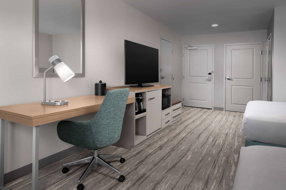 A hotel room with a wooden desk, a teal office chair, a flat-screen TV, a coffee machine, a mini-fridge, a bed with white bedding, and two white doors.