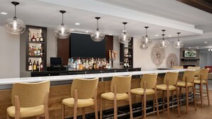 A bar area with multiple bar stools, a counter with a variety of bottles, and hanging pendant lights. - Hilton Garden Inn Marina Del Rey (Marina del Rey)