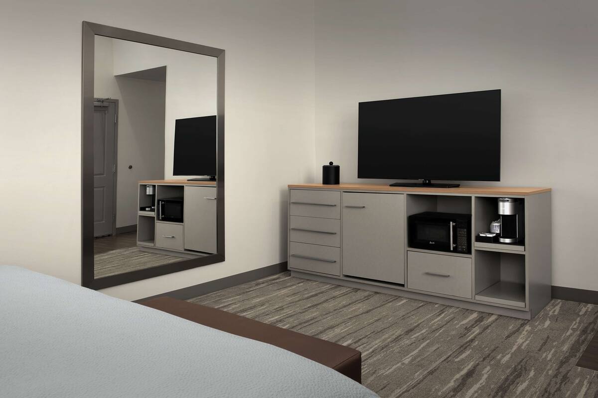 A hotel room with a bed, a large mirror, a TV, a minibar, and a coffee machine.