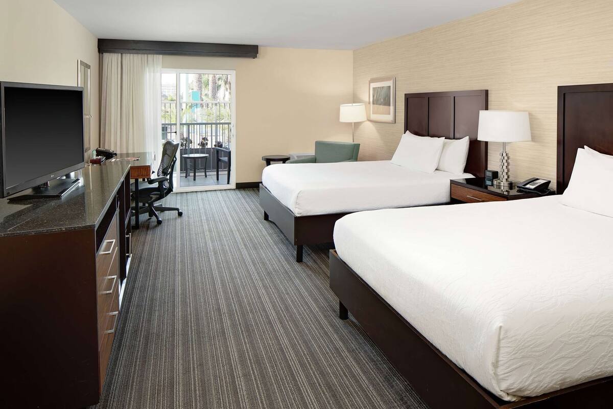 A hotel room with two beds, a desk with a TV, a chair, a lamp, and a view of the outdoors.