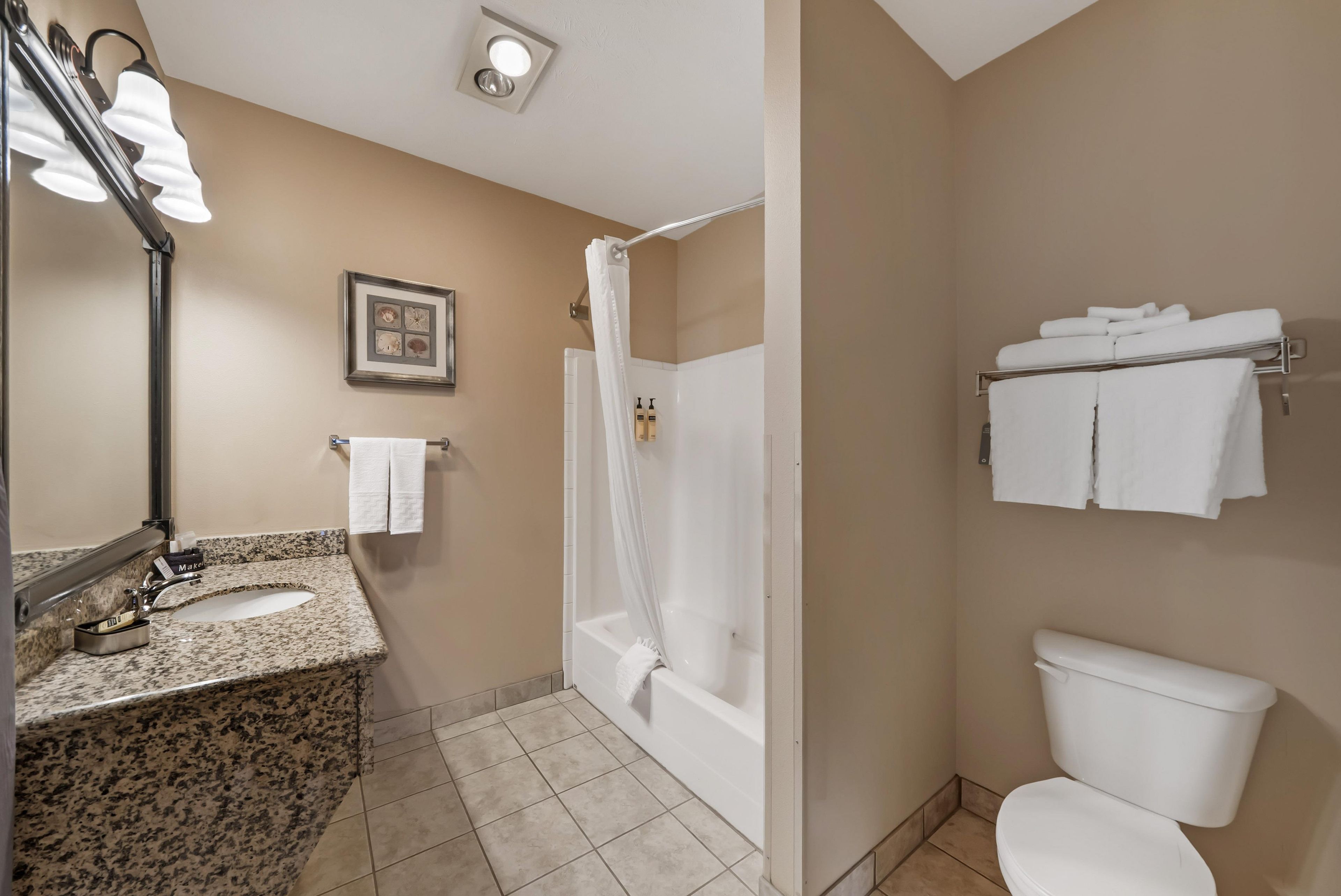A bathroom with a granite countertop, a white sink, a mirror, a framed picture on the wall, a shower curtain, a toilet, and a towel rack with towels.