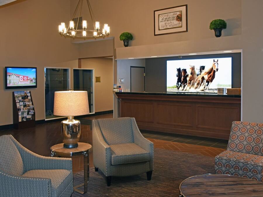 A hotel lobby with a reception desk, two armchairs, a lamp, a television displaying horses, and a framed quote on the wall.