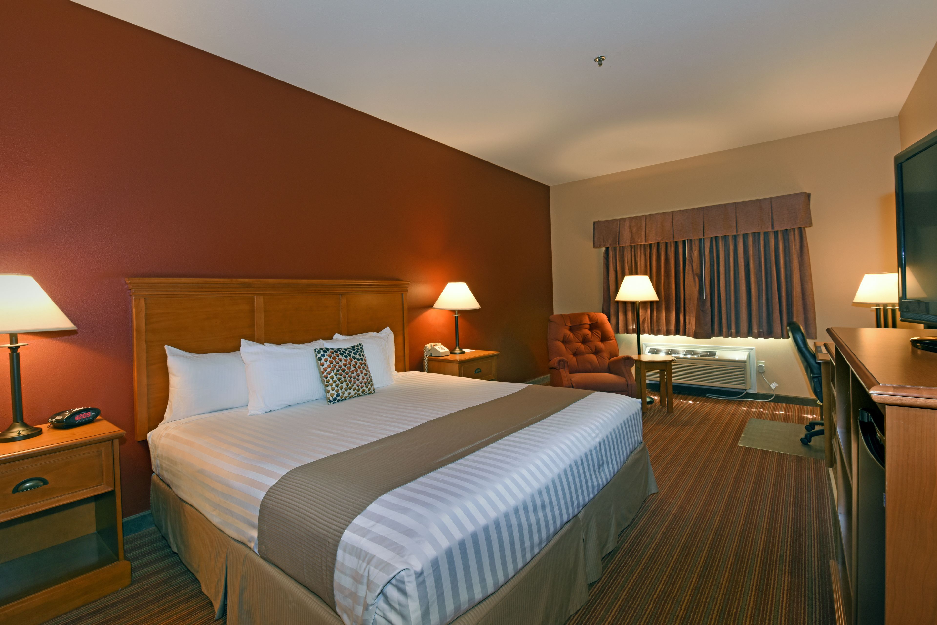 Ein Hotelzimmer mit einem großen Bett, zwei Nachttischlampen, einem Schreibtisch mit Stuhl, einem Fernseher und einem Fenster mit Vorhängen.