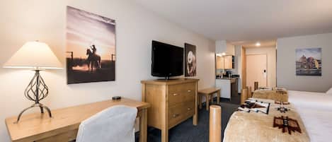 A hotel room with a desk, chair, bed, TV, and artwork on the walls.