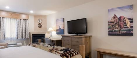 A hotel room with a large bed, a television, a desk with a lamp, a fireplace, and artwork on the walls.