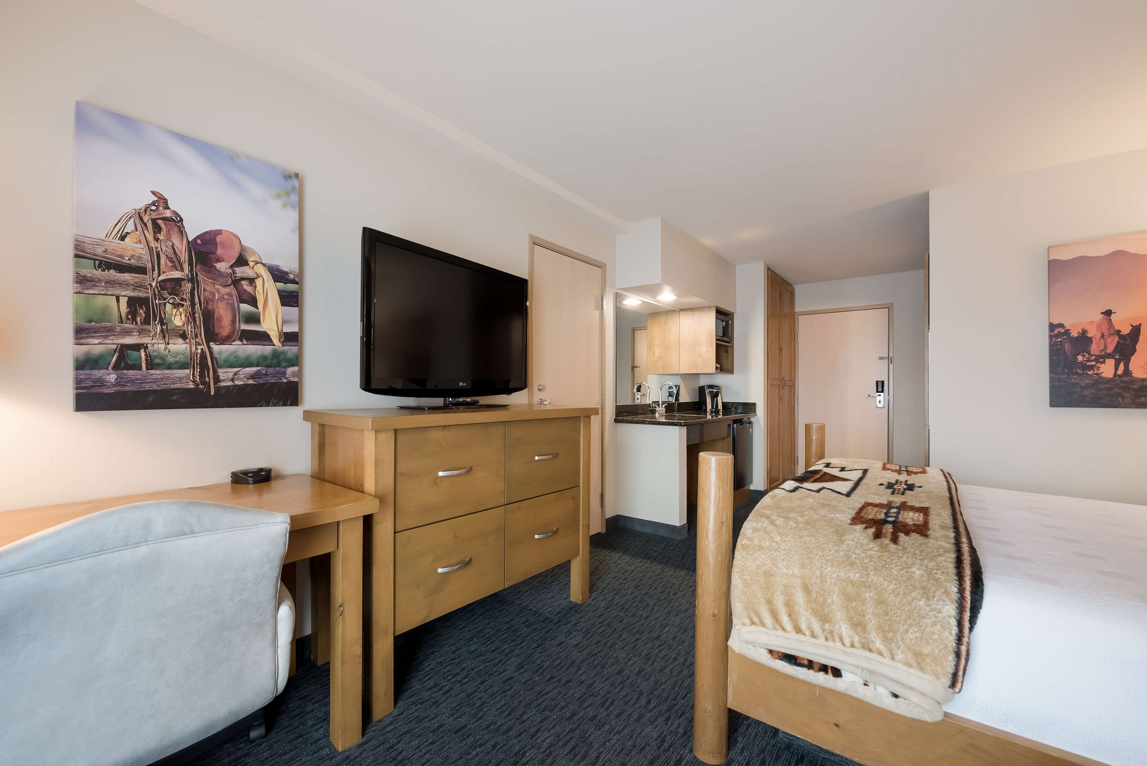 A hotel room with a bed, a desk, a television, a small kitchenette, and two paintings on the walls.
