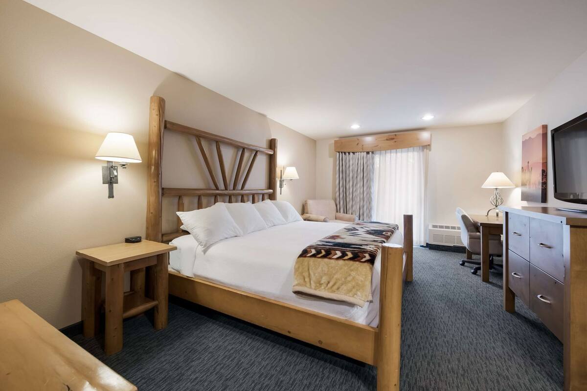 Une chambre d’hôtel avec un lit en bois, deux tables de chevet, un bureau avec une chaise, une télévision et une fenêtre avec des rideaux.