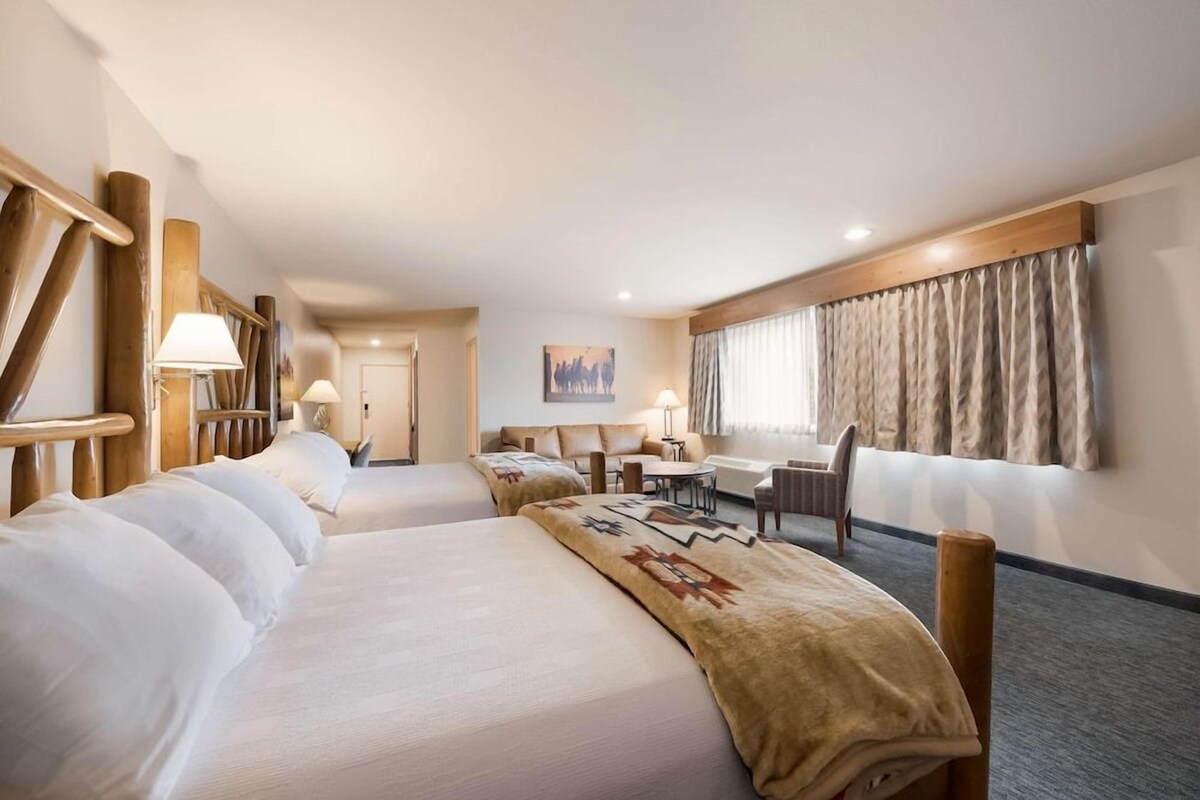 Habitación de hotel con dos camas, un escritorio, una silla, una lámpara y un ventanal con cortinas.
