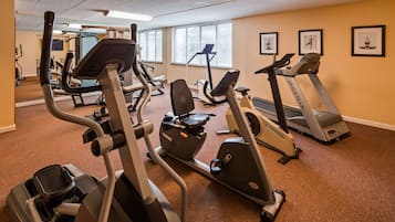 A gym with various exercise machines, including treadmills and elliptical trainers, arranged in a spacious room with carpeted flooring and large windows.