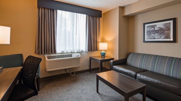 A hotel room with a sofa, a small table, a chair, a lamp, an air conditioning unit, and a window with curtains. - Best Western East Towne Suites (Madison)