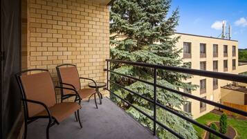 A balcony with two chairs and a small table, overlooking a brick building and a large evergreen tree.