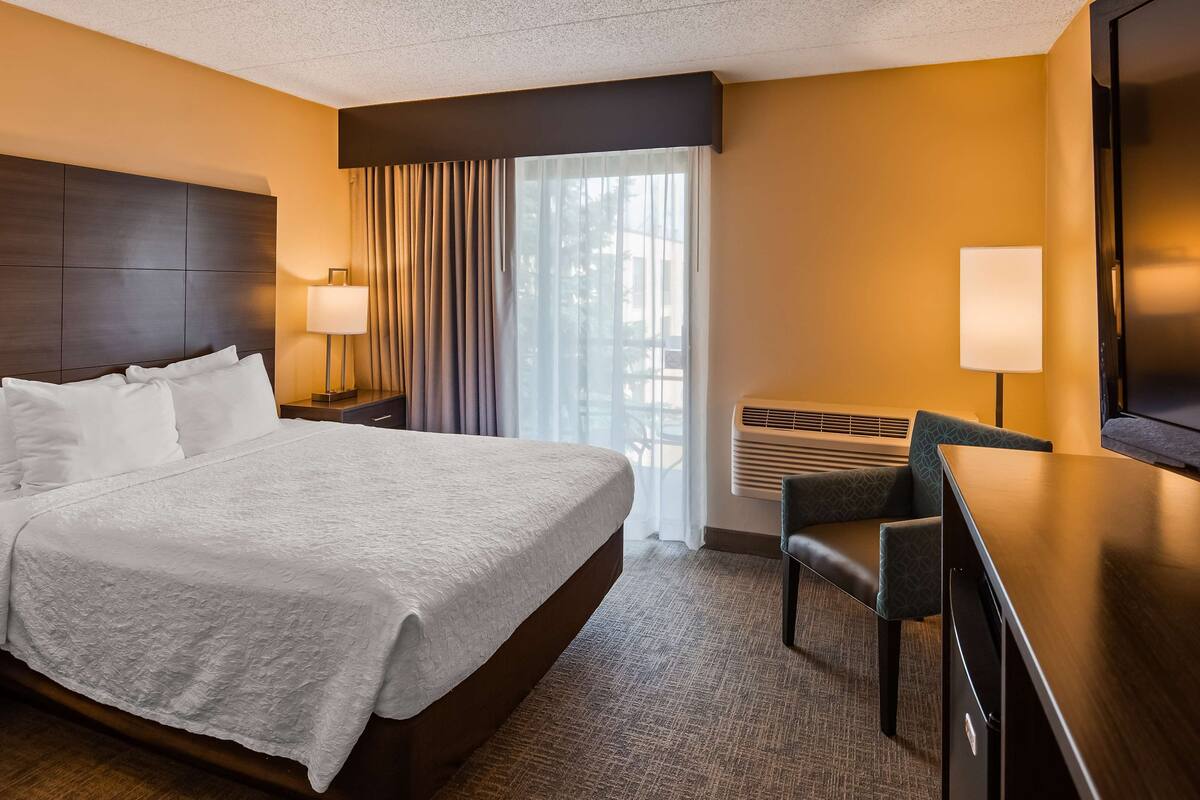A hotel room with a large bed, a desk with a chair, a flat-screen TV, a window with curtains, and a wall-mounted air conditioner.