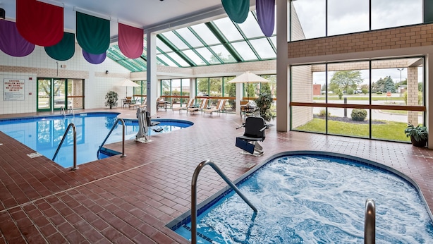 Ein Innenpool mit zwei Bereichen, einer mit klarem Wasser und der andere mit einer Whirlpool-Funktion. Um den Poolbereich befinden sich Liegestühle und Sonnenschirme, und große Fenster bieten Ausblicke nach draußen.