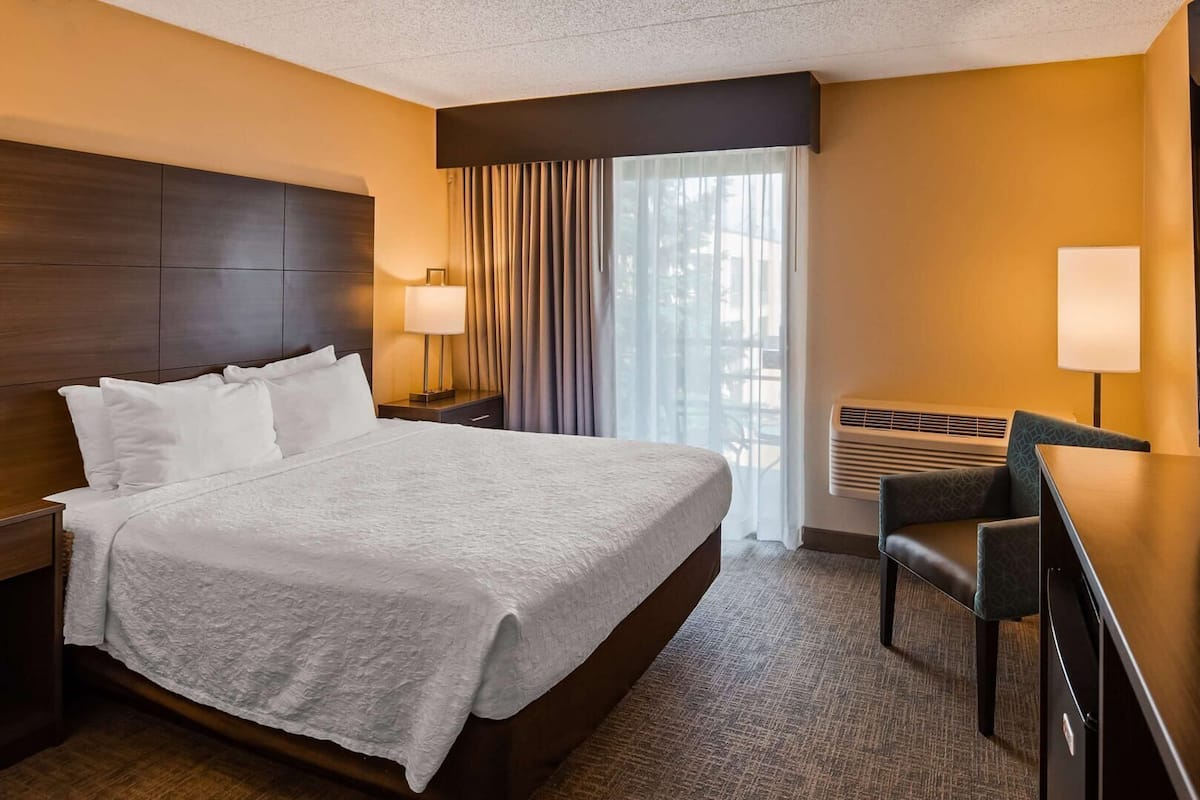 A hotel room with a large bed, a desk with a chair, a TV, a window with curtains, and a wall-mounted heater.