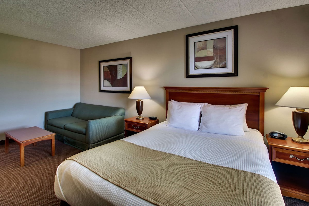 A hotel room with a bed, a green armchair, a small table, two bedside lamps, and two framed pictures on the walls.