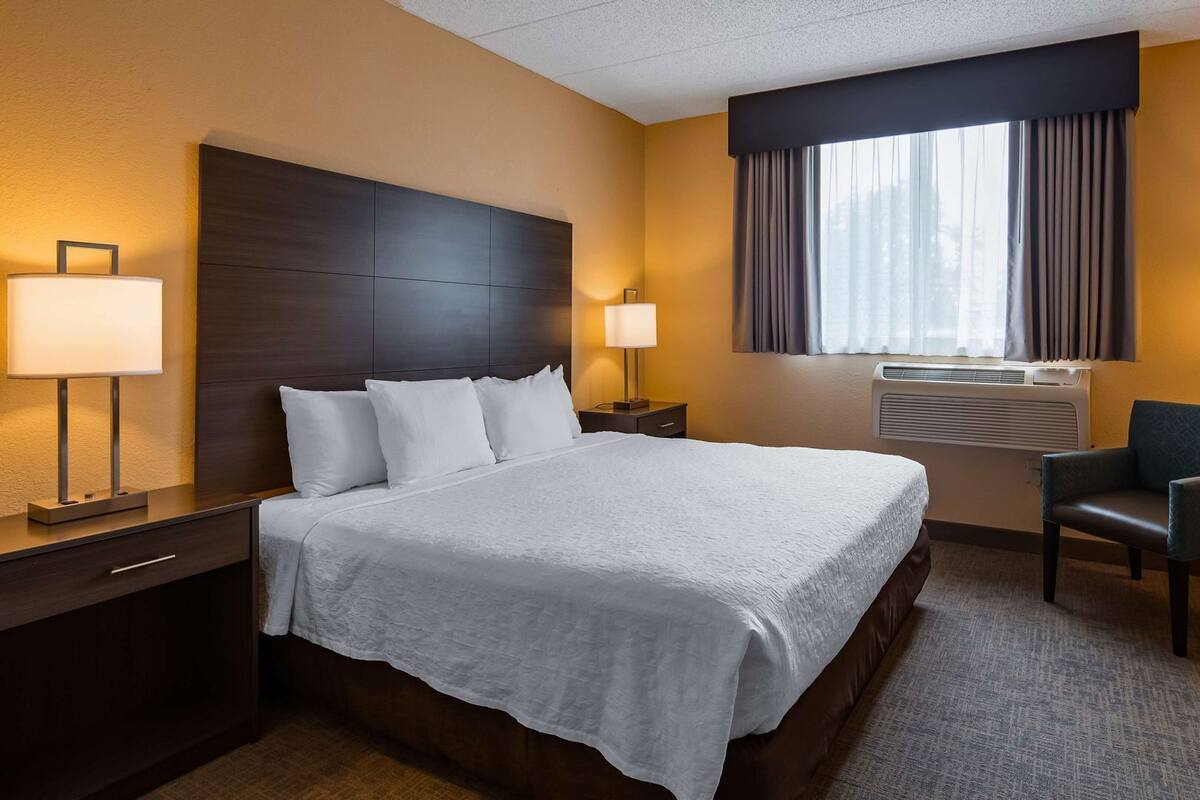 A hotel room with a large bed, two bedside tables, a chair, an air conditioning unit, and a window with curtains.
