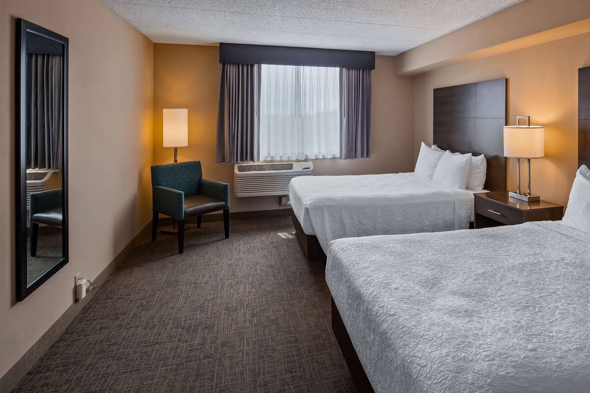 A hotel room with two beds, a chair, a desk, a lamp, and a large window with curtains.