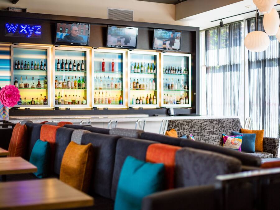 A bar area with multiple refrigerators stocked with bottles, a neon sign, and a seating area with colorful cushions.