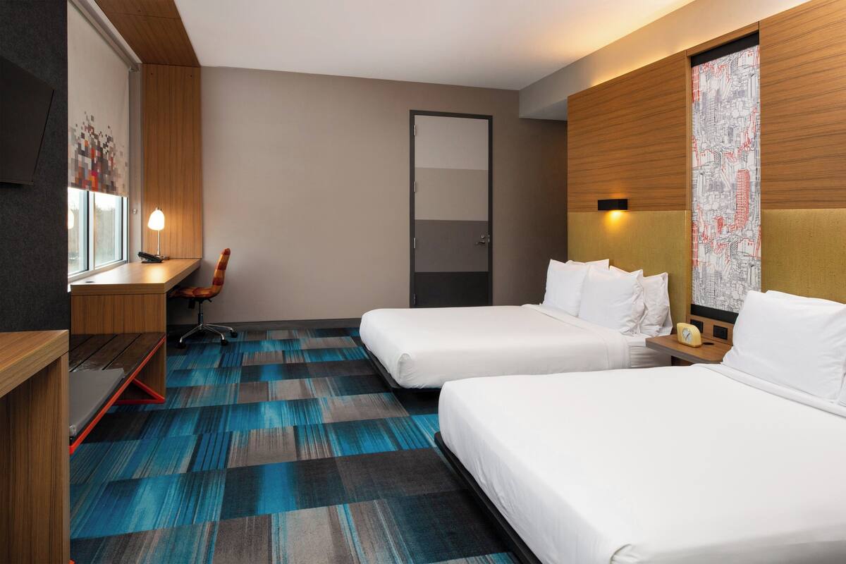 A hotel room with two beds, a desk, a chair, and a large window with blinds.