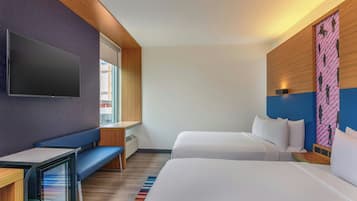 A hotel room with a large flat-screen TV, a desk, two beds, a bench, and a colorful wall decoration.