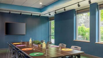 A conference room with a long table, chairs, a projector screen, and windows with a view of greenery.