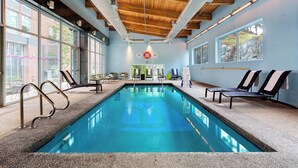 An indoor swimming pool with clear glass walls, wooden ceiling beams, and lounge chairs along the sides. - Aloft by Marriott Lexington (Lexington)