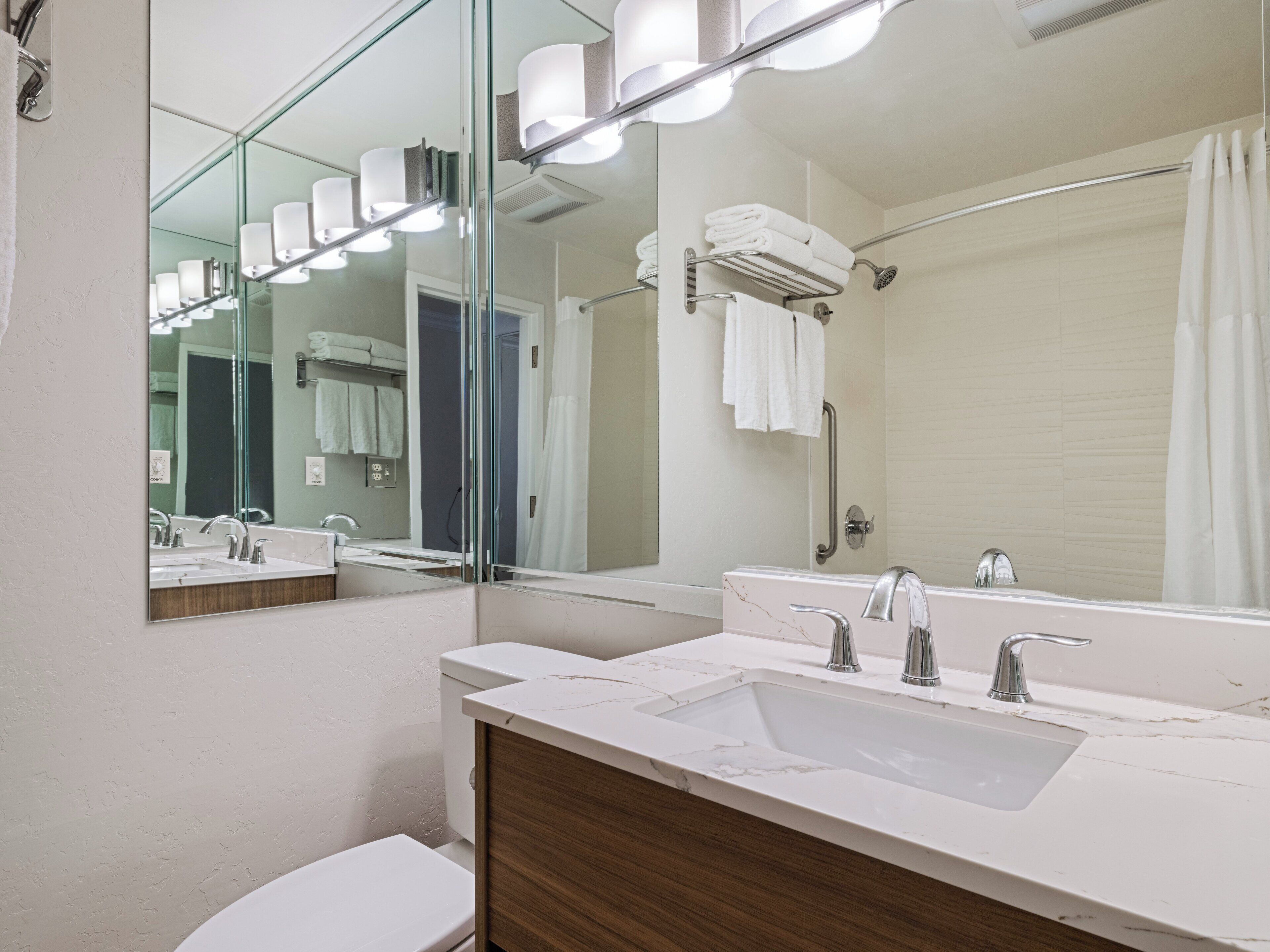 A modern bathroom with a large mirror, two sinks, a shower, and a toilet.
