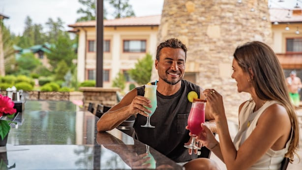 A couple sitting at an outdoor bar with drinks, overlooking a stone building and greenery.