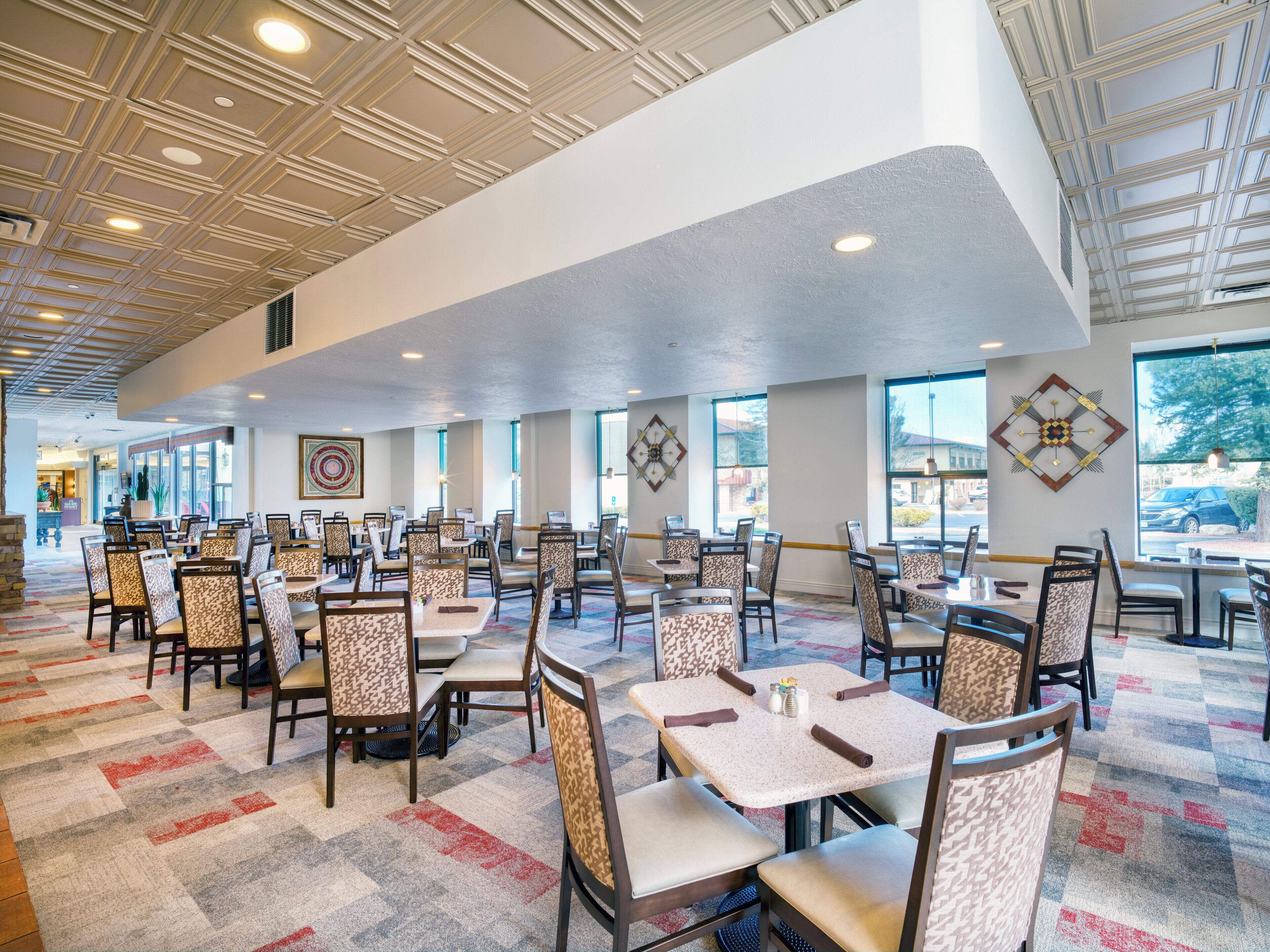 A spacious dining area with multiple tables and chairs, large windows, and a patterned carpet.