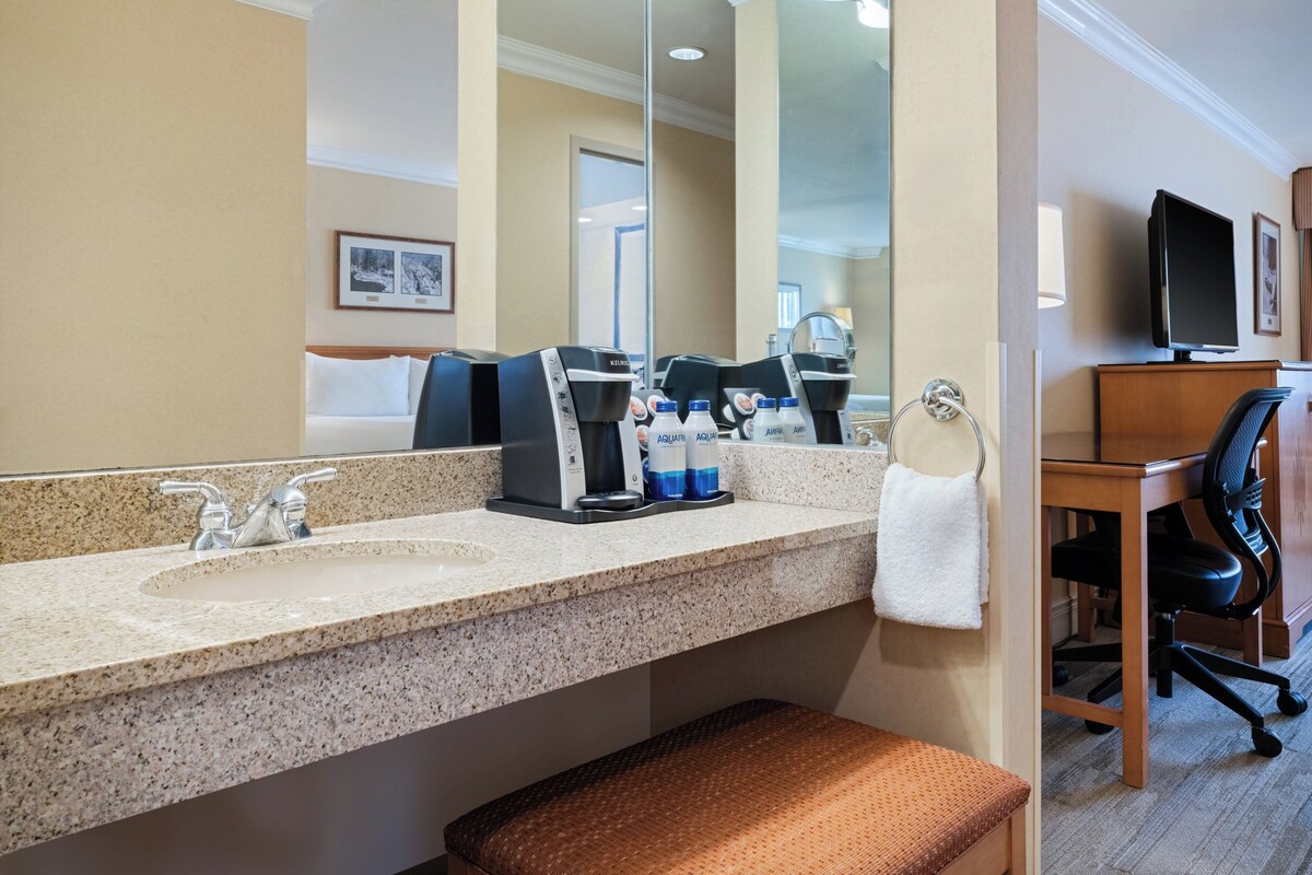 Un baño de hotel con encimera de granito, una cafetera, dos botellas de agua, una toalla, un lavabo, un espejo y un televisor sobre un escritorio de madera.
