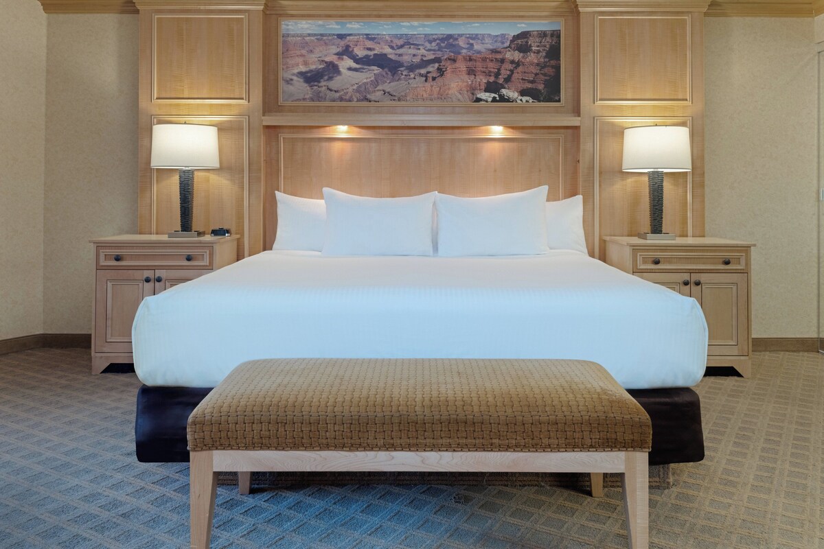 Habitación de hotel con una cama grande, dos mesitas de noche con lámparas, un banco y un cuadro colgado en la pared que muestra un cañón.