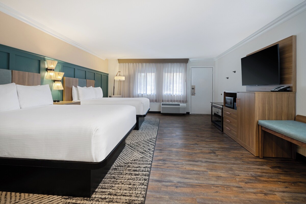 Habitación de hotel con dos camas, televisión, escritorio y un ventanal grande con cortinas.
