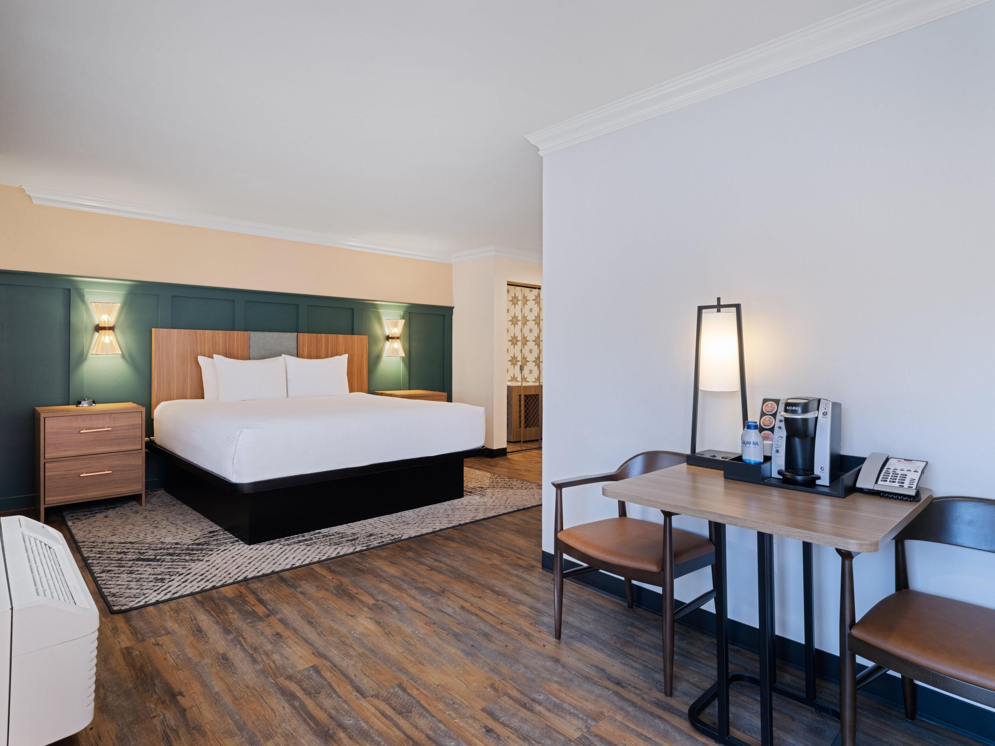 Hotelkamer met een bed, nachtkastje, lamp, bureau, stoel en een koffiezetapparaat.