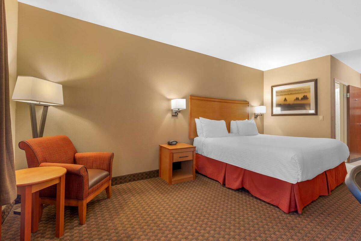 A hotel room with a bed, a chair, a nightstand, a lamp, and a painting on the wall.
