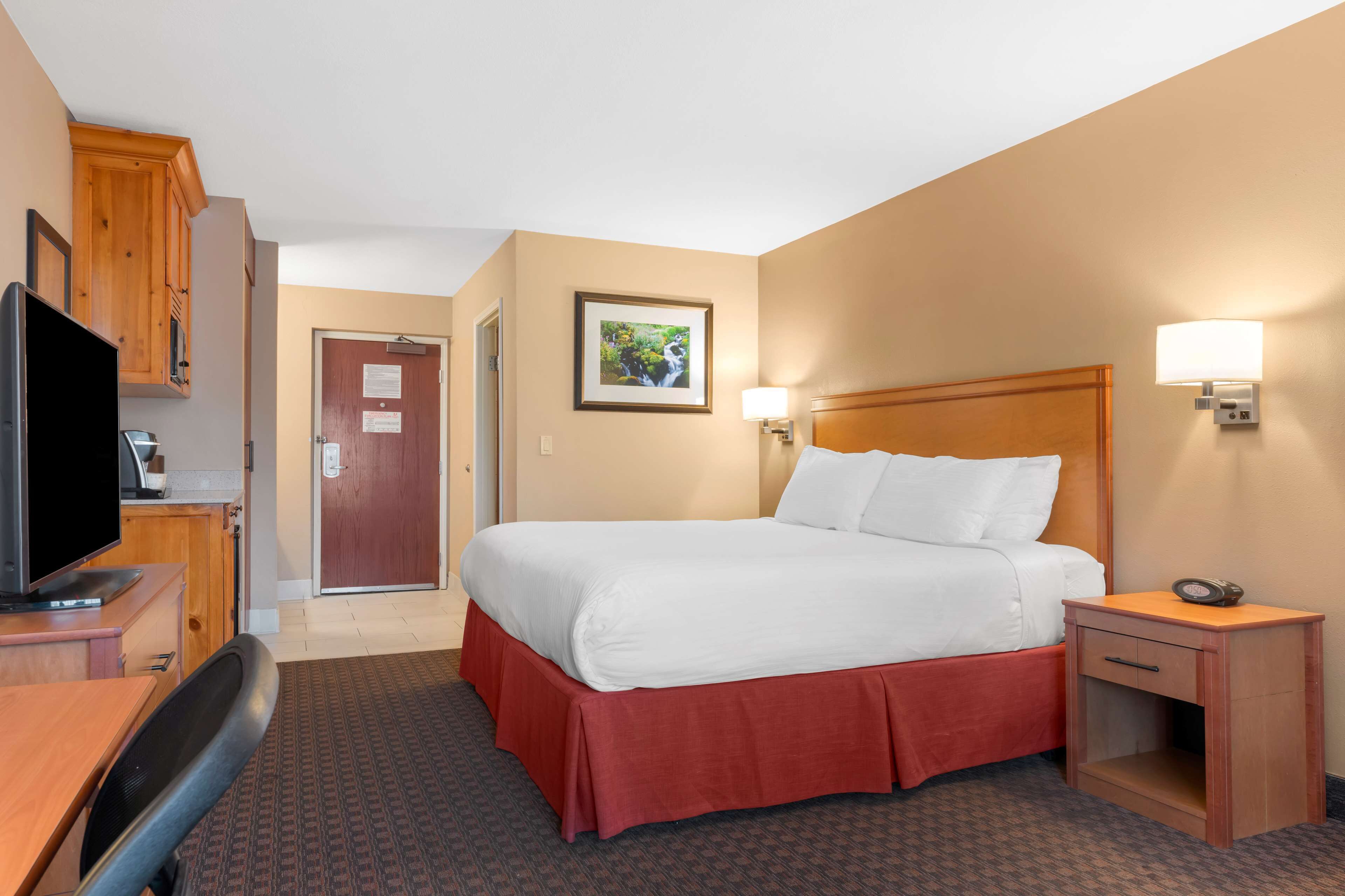 A hotel room with a bed, a desk with a television, a nightstand, and a door leading to another room.