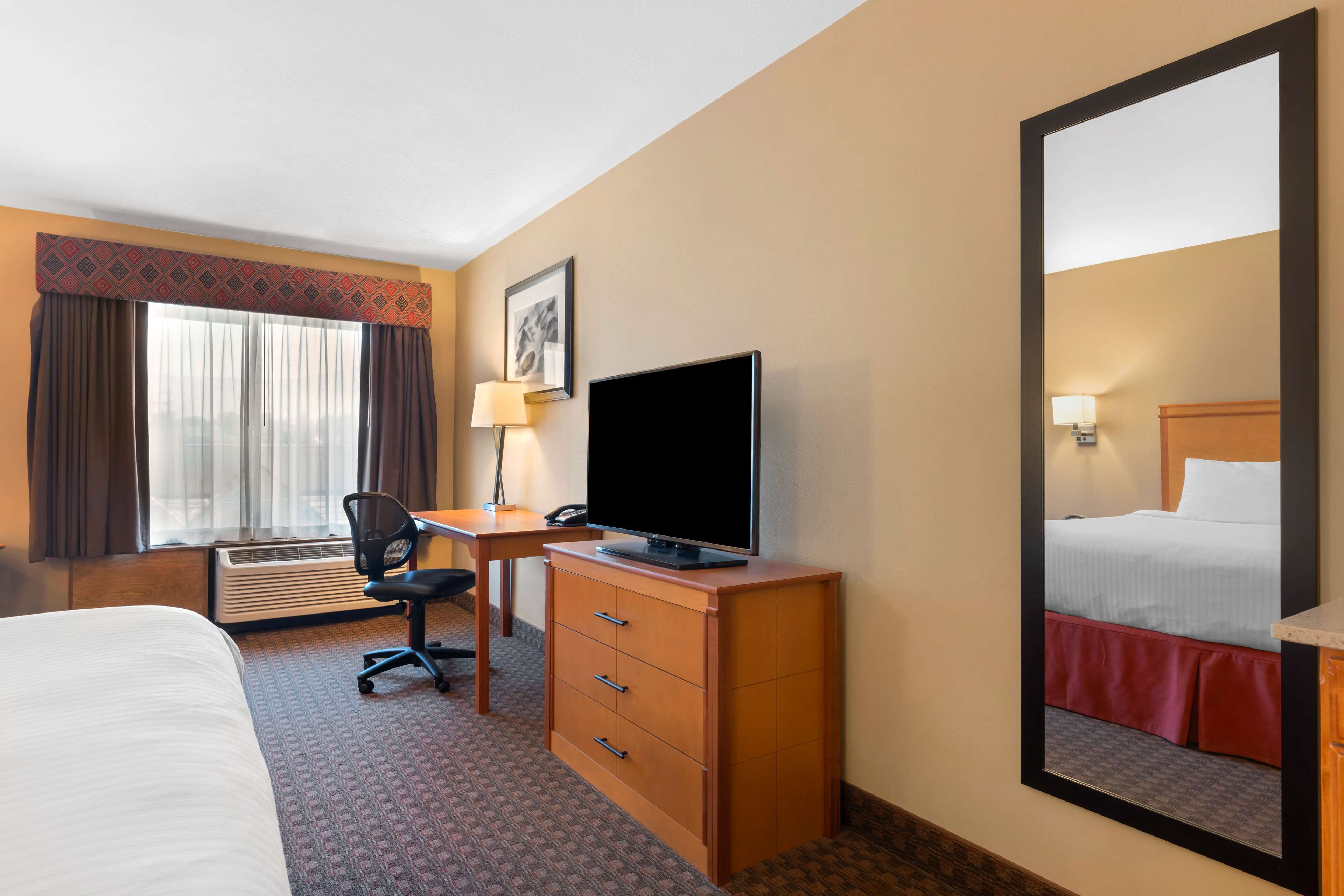 A hotel room with a bed, a desk with a television, a chair, a desk lamp, and a large mirror.
