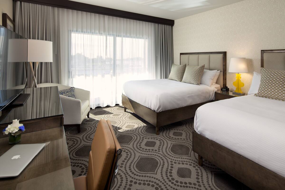 A hotel room with two beds, a desk with a laptop, a chair, a TV, and a large window with curtains.