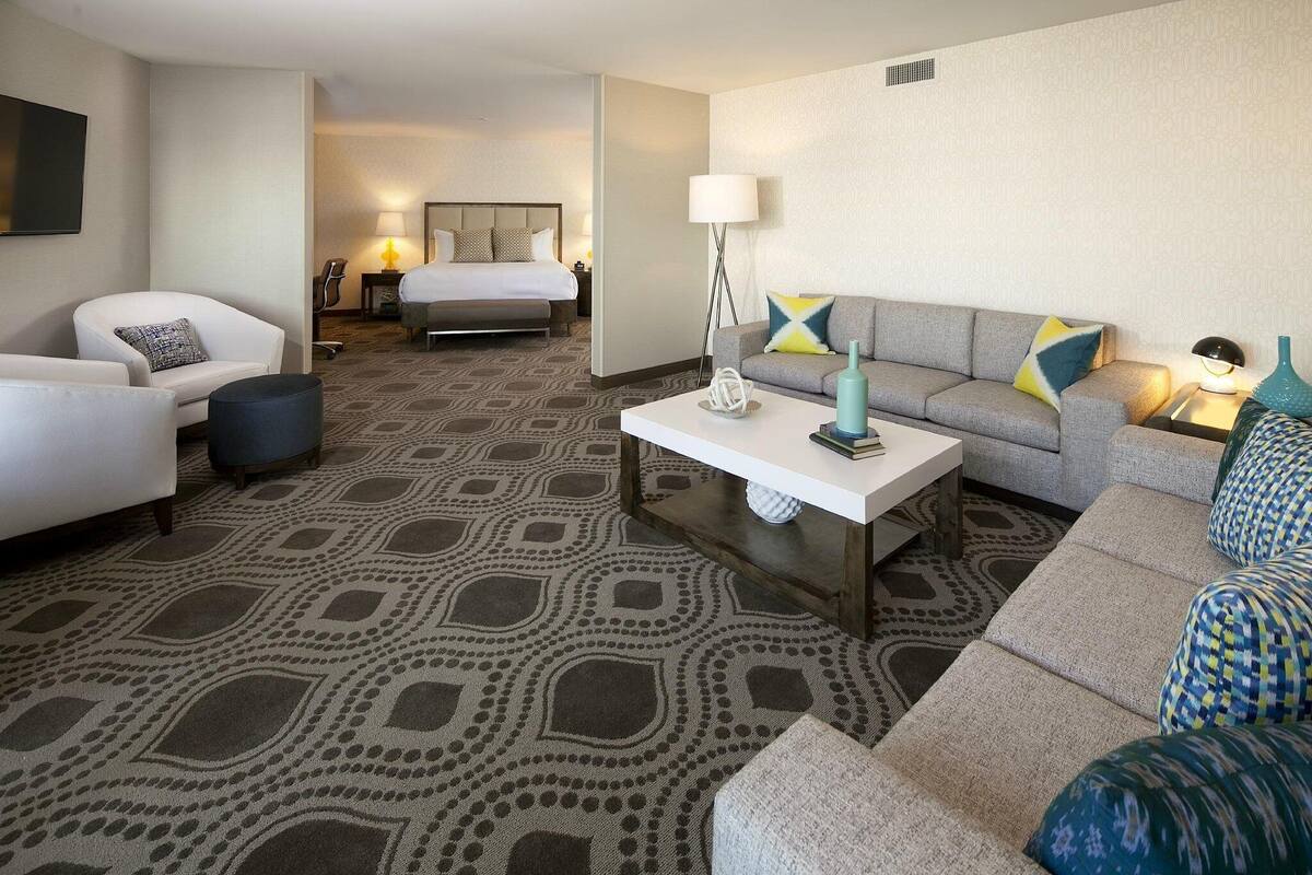 A hotel room with a patterned carpet, a sofa, a chair, a bed, and a coffee table.