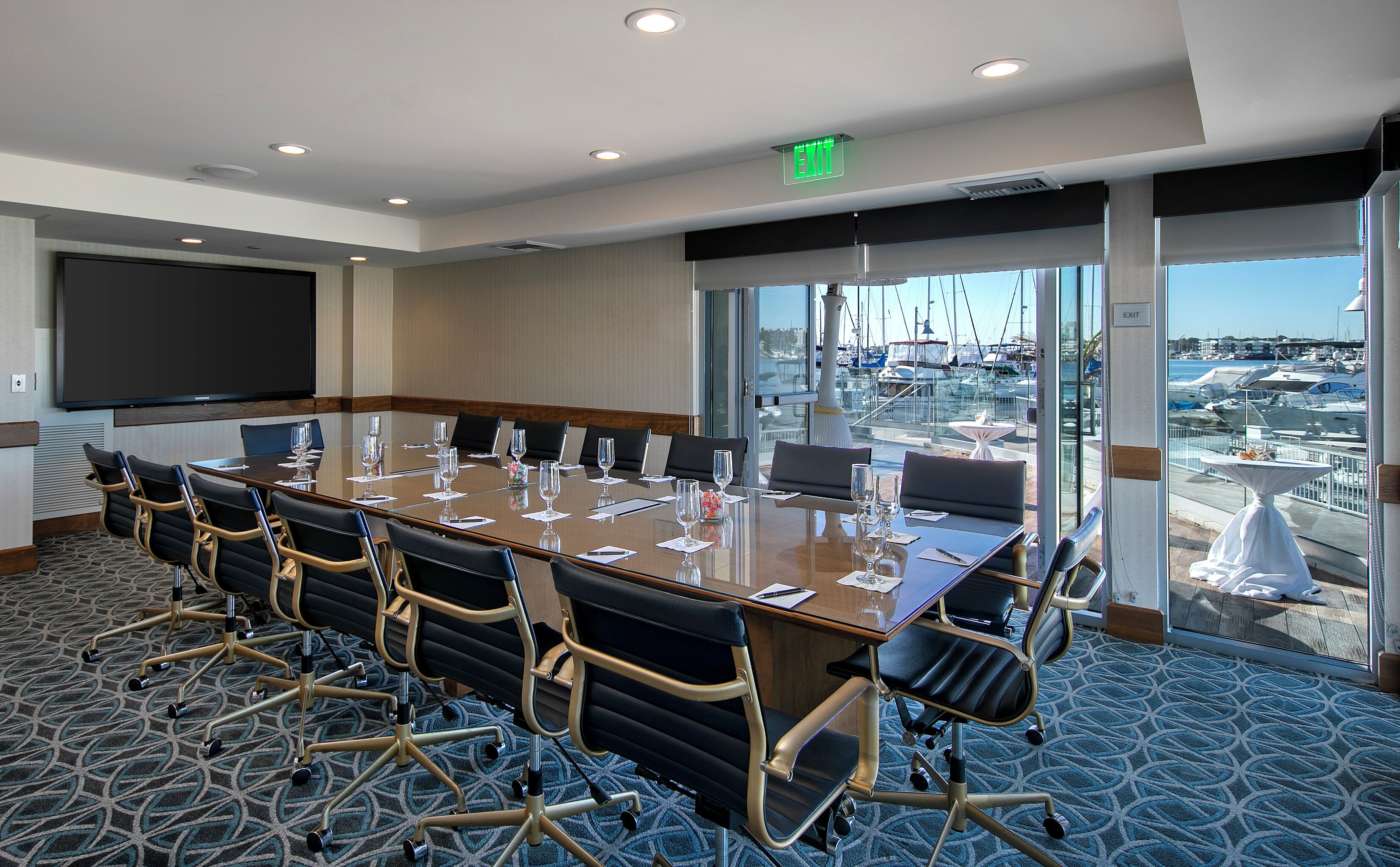 A conference room with a long table, chairs, a projector screen, and a view of a marina through large windows.