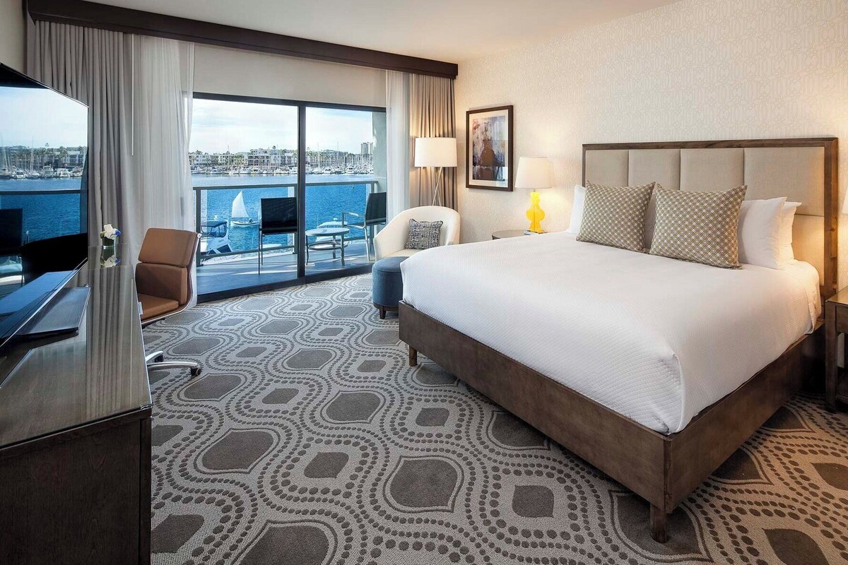 A hotel room with a large bed, a desk, a chair, a TV, and a view of the marina.