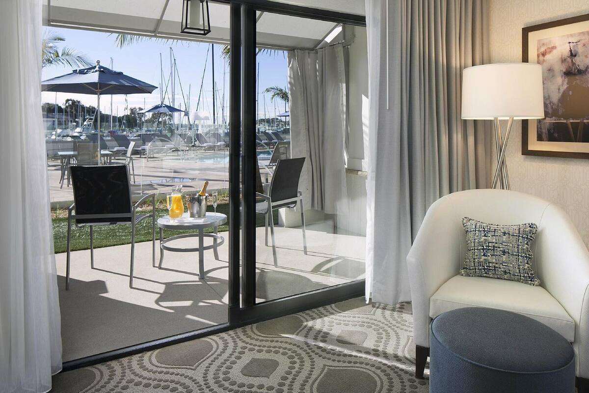 A hotel room with a glass door leading to a balcony with a view of boats and a pool. There is a white armchair and a small ottoman on the balcony.
