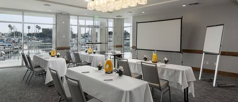 Un salón de conferencias con mesas, sillas, una pantalla de proyector y vistas al puerto deportivo.