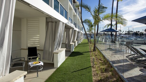 Une vue de l’extérieur d’un hôtel, avec un espace extérieur abrité aménagé en coin salon, une petite table sur laquelle est disposée une assiette de fruits, et une vue dégagée sur le port de plaisance en arrière-plan.
