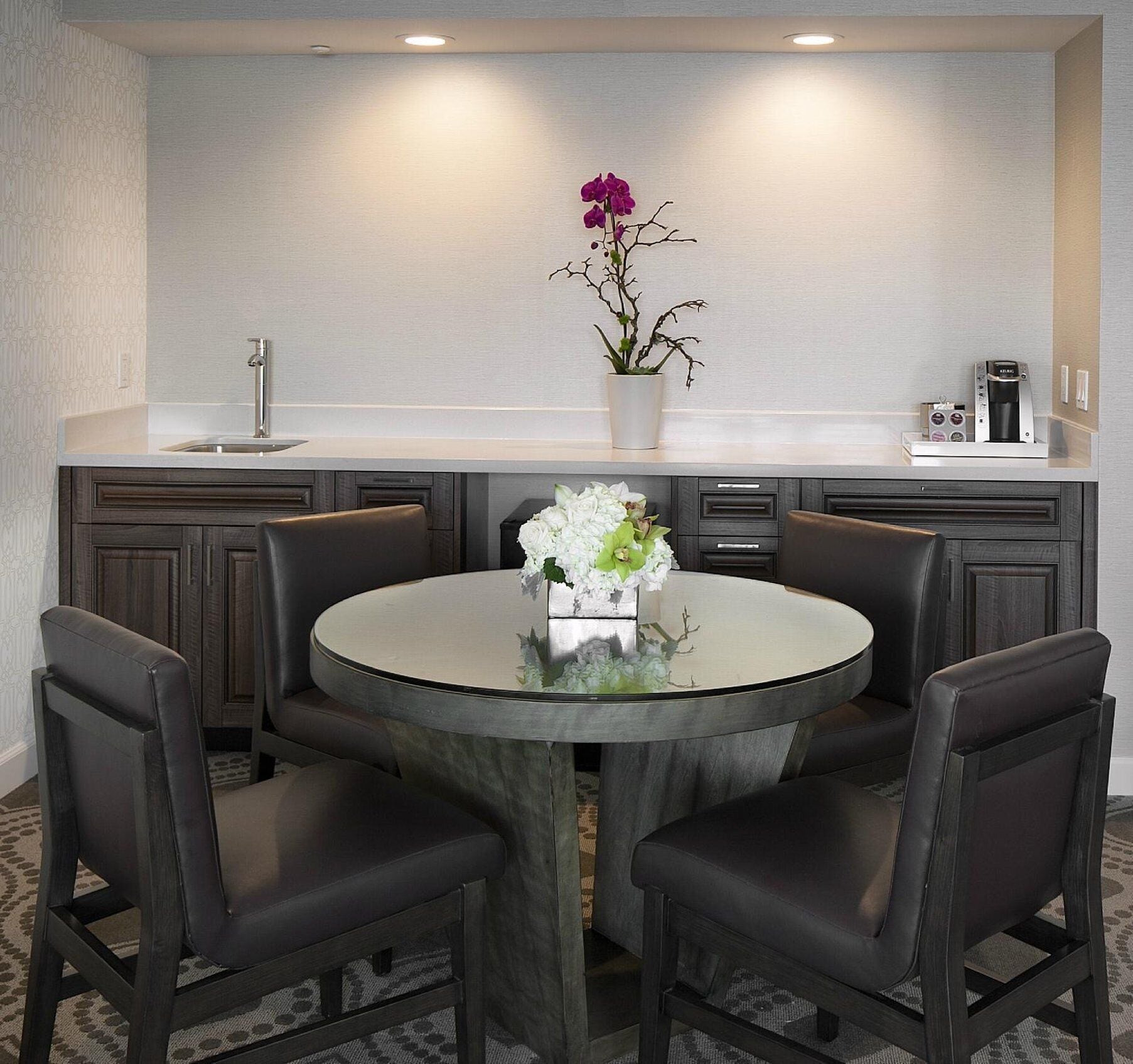 A modern kitchen with a round glass table, dark wooden cabinets, and a coffee machine.