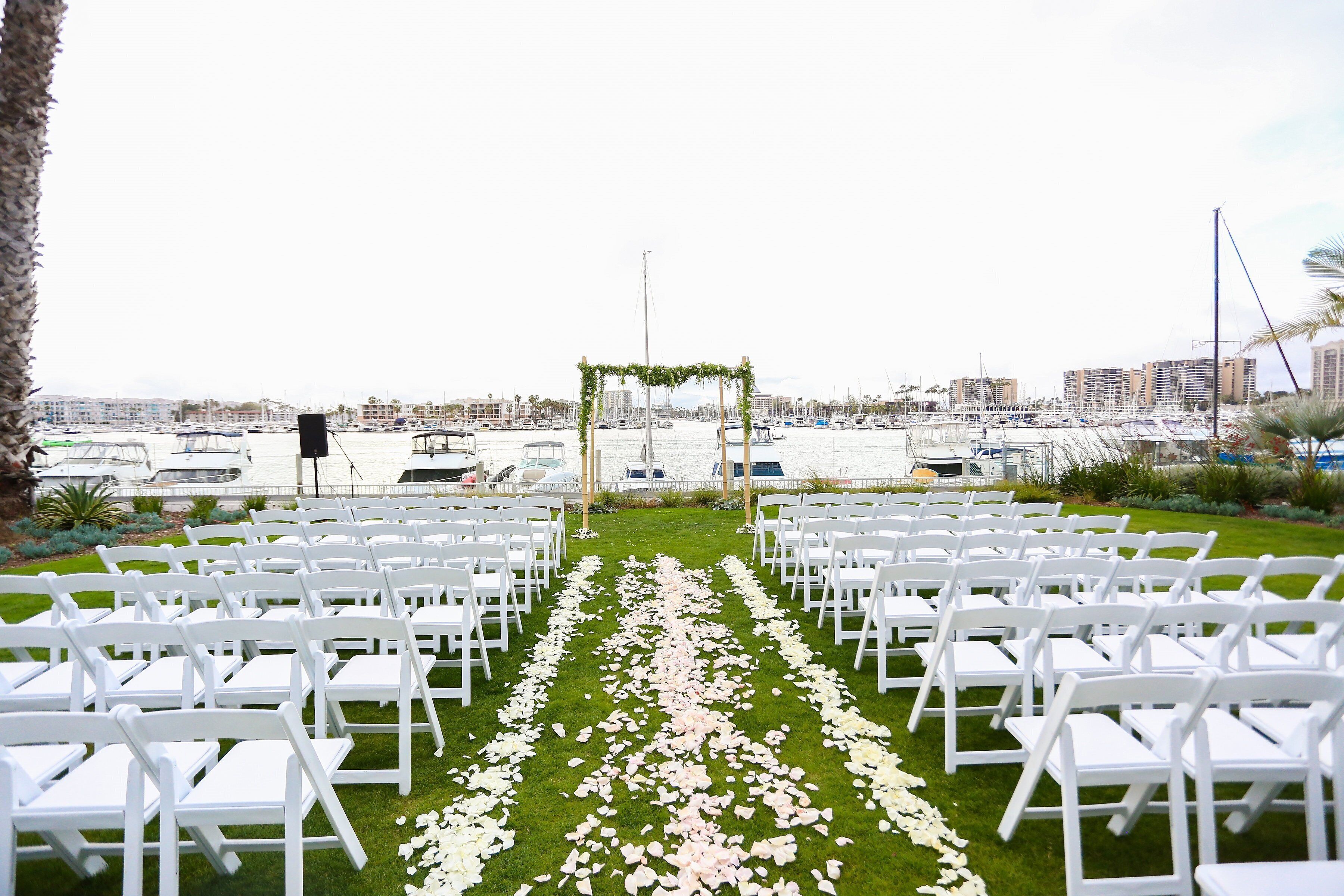 A wedding ceremony setup with white chairs and a floral aisle on a grassy area, overlooking a marina with boats and buildings.