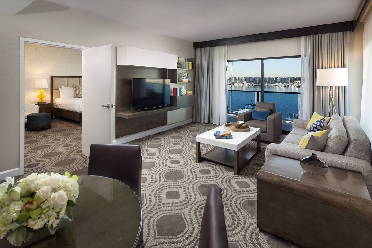 A modern hotel room with a large TV, comfortable seating, a dining area, and a view of the city.