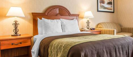A hotel room with a wooden headboard, a bed with white and brown bedding, two bedside tables with lamps, a patterned rug, and a framed picture on the wall.